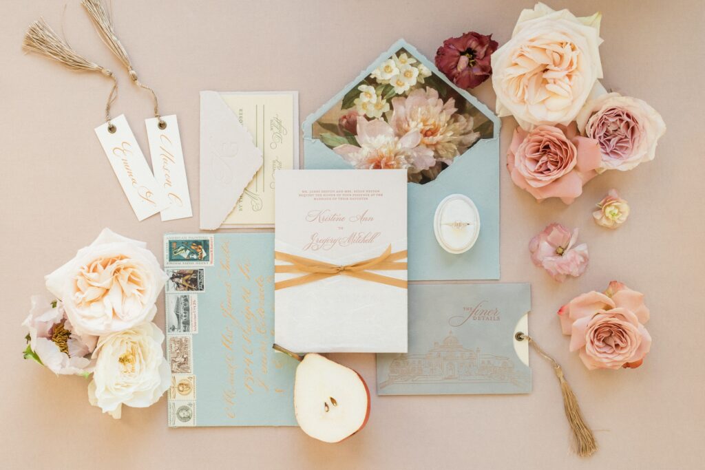 A pink and blue floral wedding invitation suite surrounded by pink peonies.